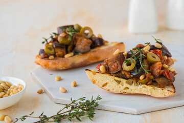 Sicilian caponata, eggplant, tomato and olive stew on white bread on a marble board against a light...