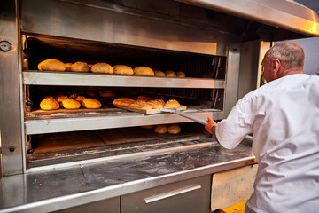 Professional baker in uniform takes out a cart with freshly baked bread from an industrial oven in a bakery