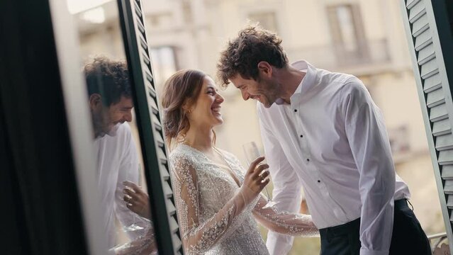 Wonderful Young Couple. Bride And Groom. Couple Drinking Champagne On Balcony Of House. View Of Magnificent Architecture. They Have Fun Talking And Kisses.