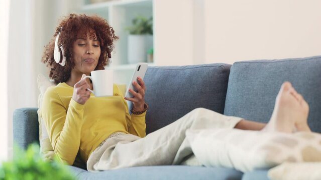 Woman taking selfies on phone, listening to music and drinking cup of coffee while relaxing on home living room sofa. Cool afro woman dancing, choosing songs from playlist and posting on social media