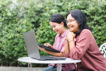 Happy senior mature and young woman playing laptop and tablet together at home garden. Asian family surfing internet with digital device with green background. Middle aged and technology