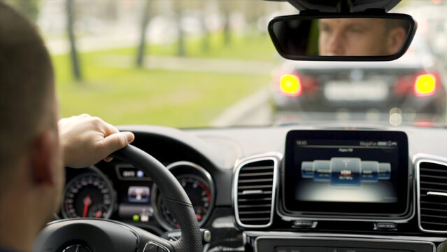 Man Driving Car From Rear View In The City Street Along Green Trees Alley. Stock. Man In Grey Shirt With Black Watch Driving Car On The Road, Modern Car Interior.