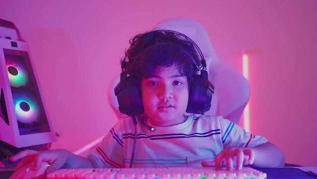 Front view of little curly-haired Asian boy in a headset staring at a screen with hands pressed on keyboard and mouse while playing online games on computer and raising fist up after winning the game.