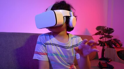 Cute little curly-haired Asian boy wearing a virtual reality headset playing game by moving his hands and gesturing touching something in the air while sitting on the couch in violet neon room at home
