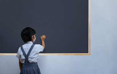 Indian school girl writing on black board while wearing protective face mask, rear view