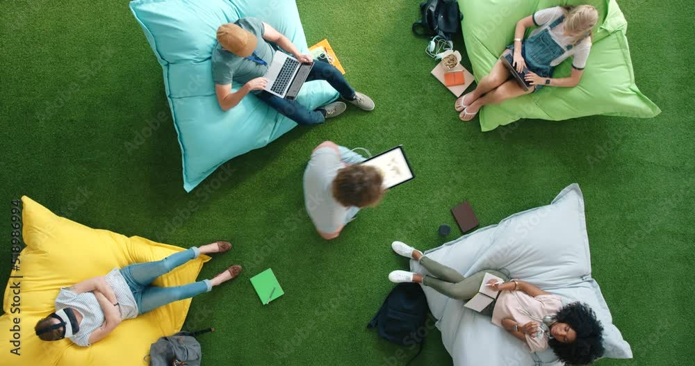 Aufkleber Aerial view of designers using smart technology while relaxing on bean bags in a casual and colorful office workspace. Student riding a hoverboard in a trendy, modern and bright library from above.