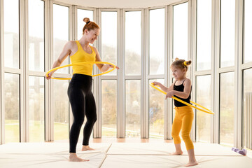 Young fit mom and her daughter having fun with hula hoop in a gym - obrazy, fototapety, plakaty