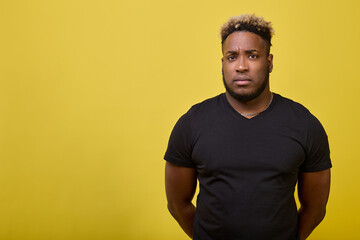 Calm black guy in a black T-shirt looks at the camera. An African-American man with a serious expression put his hands behind his back on a yellow background