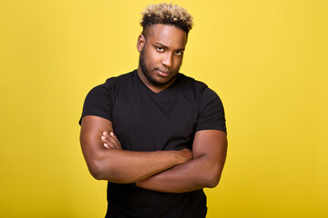 Strong guy with big muscles stands in a T-shirt on a yellow background and is sad. A disgruntled serious black man stands in a closed position, with his hands folded on his chest cross to cross. 