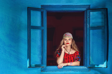 Portrait of beautiful gypsy woman in traditional red dress standing at window – Romani fortune and horoscope witch teller - 518994789