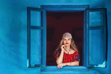 Poster Portrait of beautiful gypsy woman in traditional red dress standing at window – Romani fortune and horoscope witch teller © DanRentea