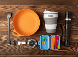 Set of tourist equipment on brown wooden background.EDC set.flat lay.Camping plate,mug,fork,flashlight and oral hygiene products.Copy space.Top view.Travel concept.Set of tourist equipment