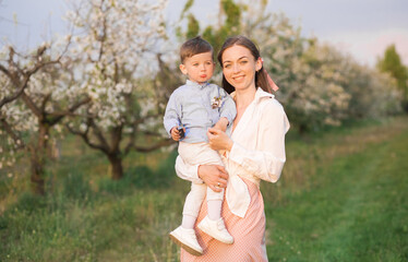 Naklejka na ściany i meble Portrait of happy mother and her baby boy. Beautiful touching scene of mom and son. Blooming garden in spring.