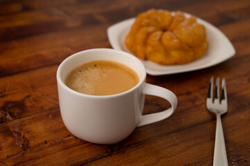 Coffee cup with round lemon cake on white plate in background and dessert fork on brown wooden table.
