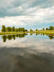 Idyllic Park and Pond Scene. Early morning views of a scenic park setting. Tranquil pond in the foreground with reflections of green leafy trees. 