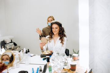 Stylist preparing bride before the wedding in a morning. The bride looks at herself in the mirror