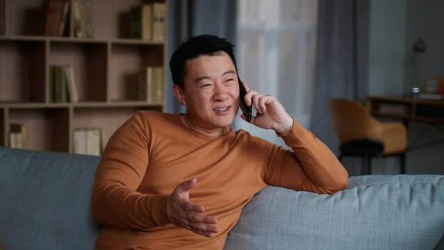 Positive middle aged asian man talking with friend via cellphone, resting on sofa at home, slow motion, free space
