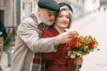 Elderly senior love couple. Old retired man woman together on romantic date.Aged husband wife walking on city street with flowers.Stylish elder hugging people pensioner in red coat.Happy family years