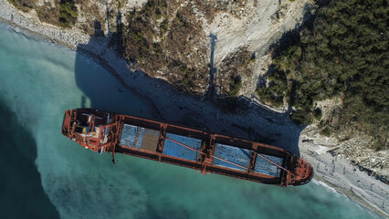 Aerial top view of red industrial ship with people on board doing sailor work moored near sea shore. Shot. Big barge on the coastline near forested slope and people walking around.