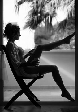 Beautiful and sexy woman sits on chair next to a balcony in bra and nightgown with a glass of drink in hand. Black and white.