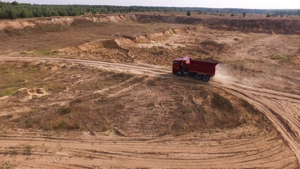 Aerial for the red big truck driving in ground quarry with flying dust behind it, industry concept. Scene. Empty lorry moving on country road with green forest on the background.