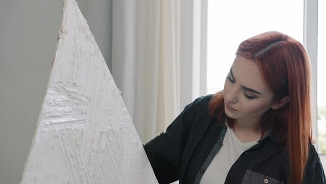 Close up red-haired Caucasian artist decorator creator designer girl 20s woman female painter create picture element of decor draws puts plaster on canvas makes painting drawing decorating modern art