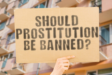 The question " Should prostitution be banned? " is on a banner in men's hands with blurred background. Labor. Lady. Law. Money. Payment. Protection. Safety. Sensuality. Prevent. Threat. Violence