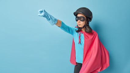 Self confident woman dressed like superhero makes flying gesture keeps arm outstretched clenches fist being on way to carnival ready to achieve goals isolated over blue background. Fictional character