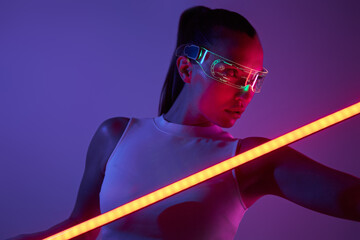 Beautiful woman in futuristic glasses holding LED lamp while standing against dark background