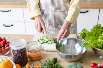 beautiful elderly gray haired senior woman cook in cozy kitchen with fresh organic vegetables, tomatoes, cabbage, lettuce, cucumbers on table cooking healthy vegetable salad, healthy food active life