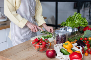 beautiful elderly gray haired senior woman cook in cozy kitchen with fresh organic vegetables, tomatoes, cabbage, lettuce, cucumbers on table cooking healthy vegetable salad, healthy food active life
