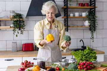 beautiful elderly gray haired senior woman cook in cozy kitchen with fresh organic vegetables,...