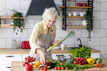 beautiful elderly gray haired senior woman cook in cozy kitchen with fresh organic vegetables, tomatoes, cabbage, lettuce, cucumbers on table cooking healthy vegetable salad, healthy food active life