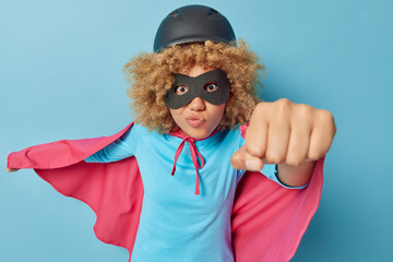 Photo of attentive curly haired female superhero makes flying gesture clenches fist dressed in masquerade costume pretends having superpower and courage poses indoor against blue background.