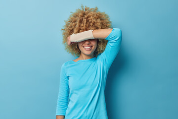 Waist up shot of happy curly haired young woman covers eyes with injured arm wears elastic bandage smiles broadly dressed in casual jumper has trauma after accident isolated over blue background