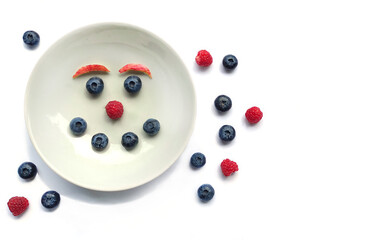Breakfast, snack for children. Funny face made of blueberries and raspberries on a white plate