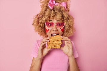 Photo of impressed woman with curly hair wears bowtie on head bites delicious appetizing waffles applies hydrogel patches under eyes to reduce puffiness wears pink sunglasses and casual t shirt