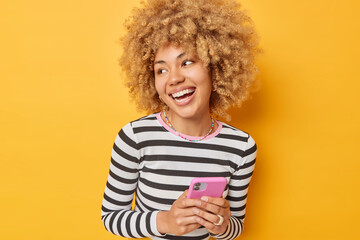 Waist up shot of positive young woman with curly hair smiles broadly holds mobile phone chats in...