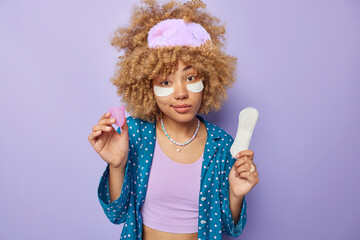 Photo of calm curly haired European woman wears sleepmask and pajama holds rubber menstrual cup and sanitary napkin isolated over purple background. Zero waste aleternative gynecology concept