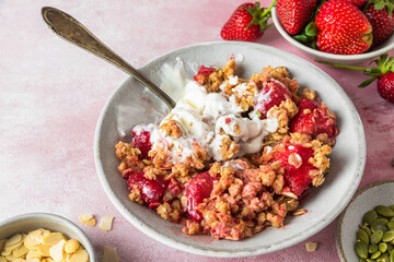 Delicious strawberry crumble with ice cream, almonds and pumpkin seeds in a plate with spoon on pink background