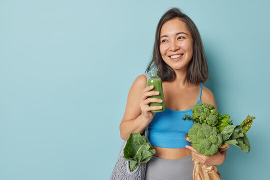 Indoor shot of cheerful dark haired Asian woman holds freshly made smoothie from vegetables carries net bag wears cropped top and leggings isolated over blue background blank space for promo