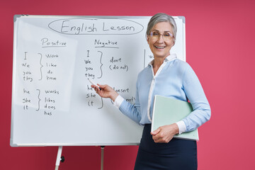 Fototapeta premium Cheerful senior woman teaching English language near the whiteboard against pink background