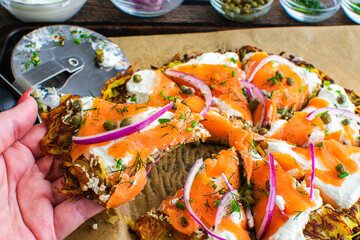 Holding a Slice of a Potato Pancake Topped with Smoked Salmon and Labneh: Latkes topped with nova, capers, red onions, and fresh herbs