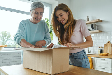 Excited senior woman and her adult daughter unpacking box while standing at the domestic kitchen