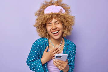 Overjoyed European woman with curly hair laughs happily keeps hand on chest uses smartphone reads...