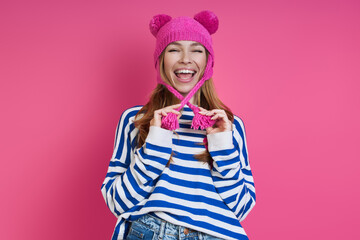 Playful young woman adjusting her pink hat and smiling while standing against colored background