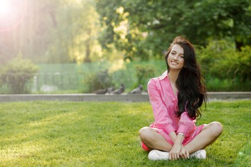 Fototapeta premium The girl in a pink dress sits on the grass with her legs crossed