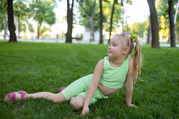 Naklejka na ściany i meble a little girl in a green suit sits on the lawn in the park and poses for the camera