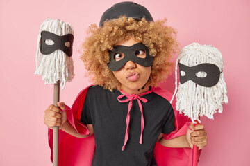 Funny young woman pretends being superhero makes grimace holds two mops wears helmet eyemask and...