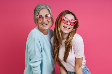 Happy mother and adult daughter wearing funky eyeglasses while standing together against pink background